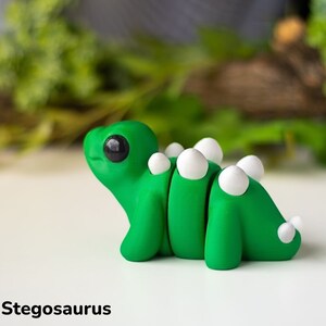 May include: A small green and white clay figurine of a baby stegosaurus. The dinosaur has a large head with big black eyes and a small mouth. It has a row of white bumps down its back.
