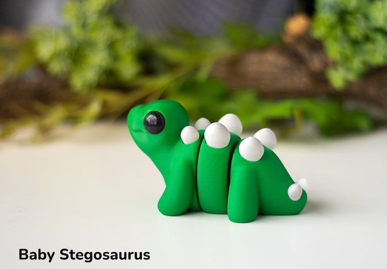 May include: A small green and white clay figurine of a baby stegosaurus. The dinosaur has a large head with big black eyes and a small mouth. It has a row of white bumps down its back.