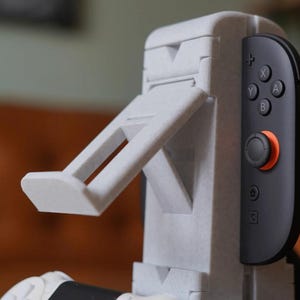 May include: A light gray 3D-printed gaming console stand with an adjustable support arm. The stand is designed to hold a black and gray game controller. The controller has orange and black accents and visible buttons.