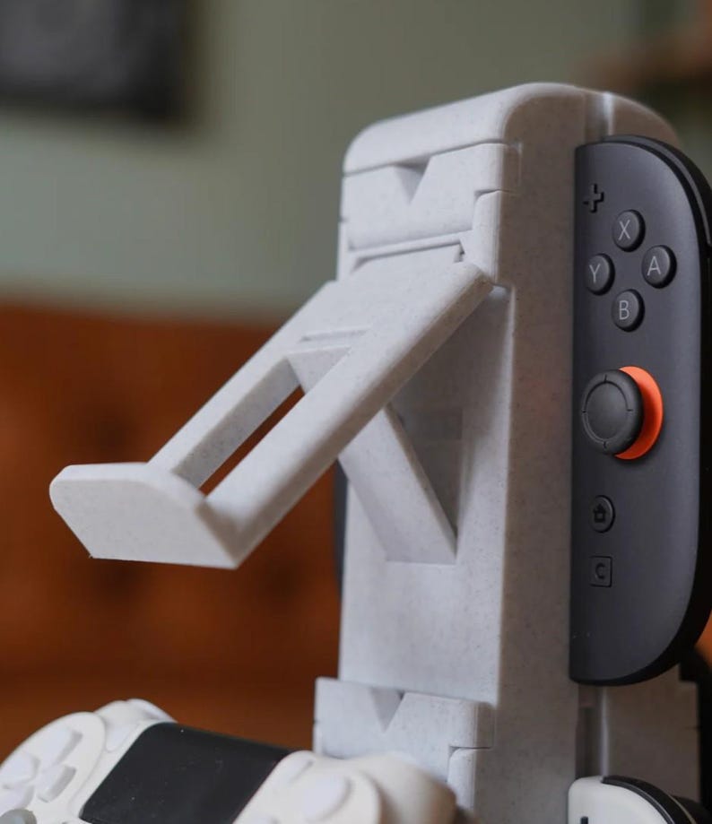 May include: A light gray 3D-printed gaming console stand with an adjustable support arm. The stand is designed to hold a black and gray game controller. The controller has orange and black accents and visible buttons.