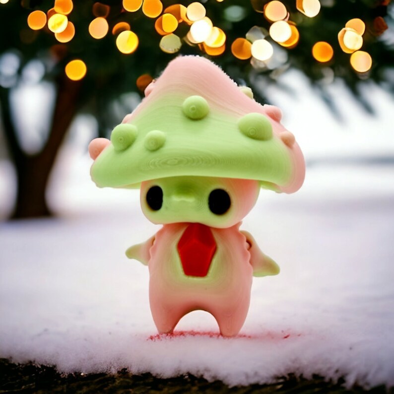 May include: A whimsical, mushroom-shaped figurine with a pink body, a green cap, and a red gem-like detail on its chest. The figurine has large black eyes and is standing in the snow. The background features a blurred Christmas tree with bokeh lights.
