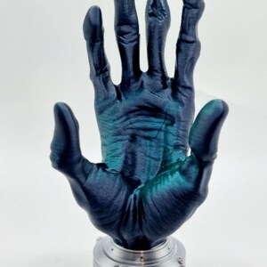 May include: A 3D printed hand sculpture with a dark blue and green metallic finish. The hand is raised with fingers spread apart, and it is mounted on a silver base.