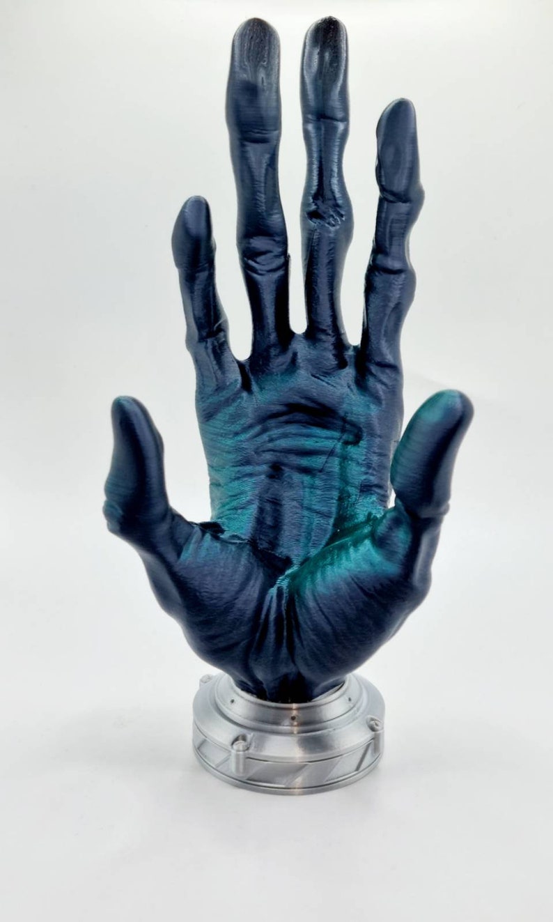 May include: A 3D printed hand sculpture with a dark blue and green metallic finish. The hand is raised with fingers spread apart, and it is mounted on a silver base.