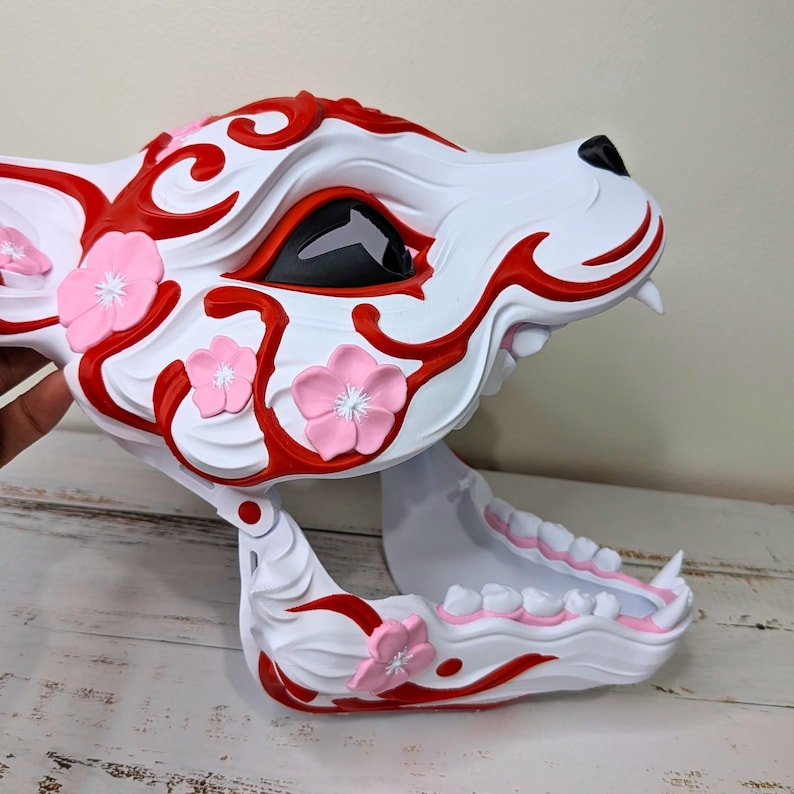 May include: A white and red decorative mask in the shape of a wolf's head, adorned with pink cherry blossoms. The mask features a detailed design with open jaws and black eye sockets.