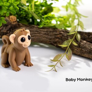 May include: A small brown toy monkey with large eyes and a curled tail. The monkey is sitting on a white surface with a branch and green leaves in the background. The text "Baby Monkey" is visible below the monkey.