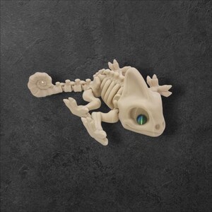 May include: A beige plastic chameleon skeleton with a green eye. The skeleton is posed in a walking position with its tail curled.