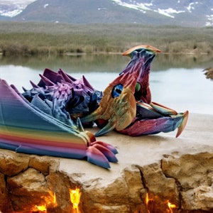 May include: A rainbow-coloured 3D printed dragon sculpture lying on a rocky surface near a lake and mountains. The dragon's wings are spread out and its head is turned to the side.