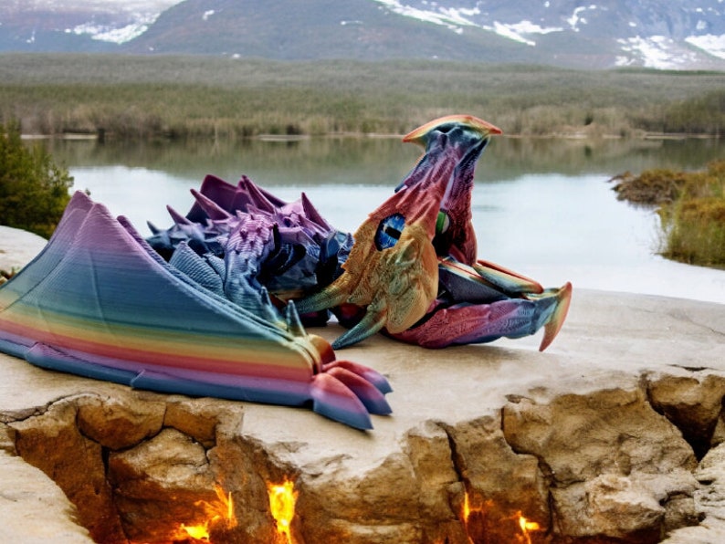 May include: A rainbow-coloured 3D printed dragon sculpture lying on a rocky surface near a lake and mountains. The dragon's wings are spread out and its head is turned to the side.