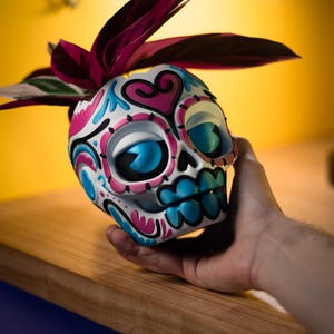 May include: A Day of the Dead skull-shaped planter, white with pink and blue accents, holds a plant with dark red leaves. The skull has large blue eyes and a detailed floral design. The planter is held in a hand.