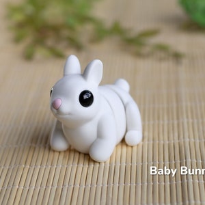 May include: A small, white ceramic bunny figurine with black eyes and a pink nose. The bunny is sitting on a woven bamboo mat.