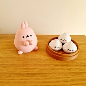 May include: A pink bunny figurine with a sad expression, next to a brown bamboo steamer with three white steamed buns with happy faces.