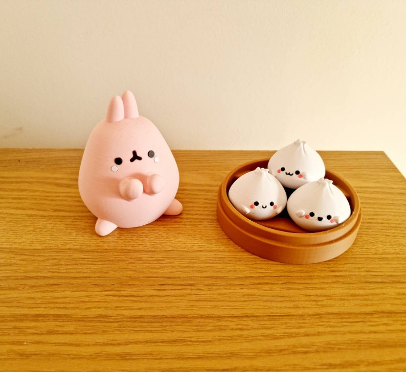 May include: A pink bunny figurine with a sad expression, next to a brown bamboo steamer with three white steamed buns with happy faces.