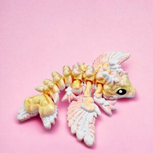 May include: A small, yellow and white dragon figurine with wings, lying on its side on a pink background.