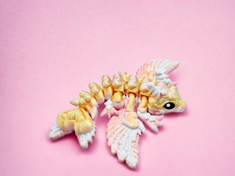 May include: A small, yellow and white dragon figurine with wings, lying on its side on a pink background.