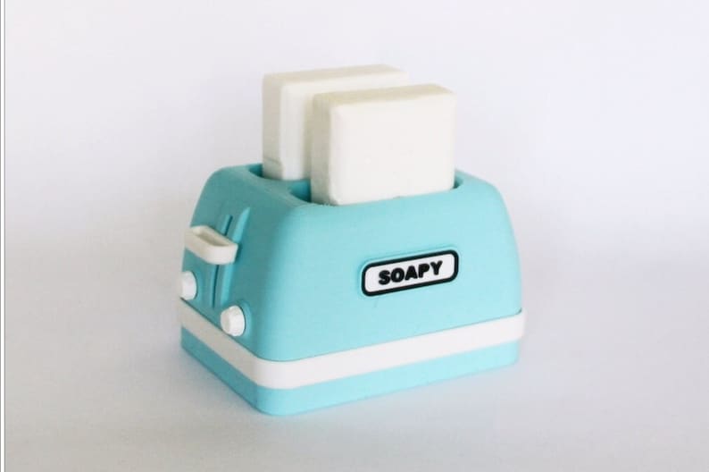 K&ouml;nnte beinhalten: Ein hellblauer Toaster mit wei&szlig;en Akzenten und dem Wort "SOAPY" auf der Vorderseite. Zwei wei&szlig;e Seifenst&uuml;cke befinden sich in den Toasterschlitzen.