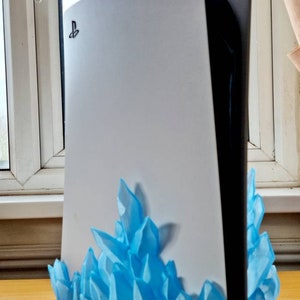 May include: A white PlayStation 5 console with a blue crystal-shaped stand. The console has the PlayStation logo on the front.