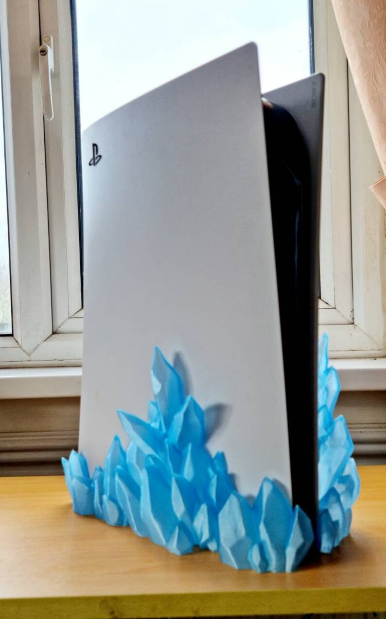 May include: A white PlayStation 5 console with a blue crystal-shaped stand. The console has the PlayStation logo on the front.