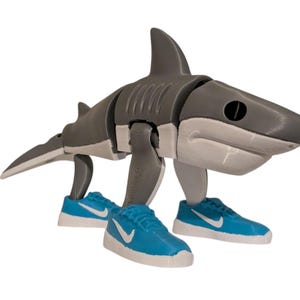 May include: A gray and white articulated shark figure wearing blue sneakers with white accents. The shark has a segmented body and a fin on its back. The sneakers have a white swoosh logo.