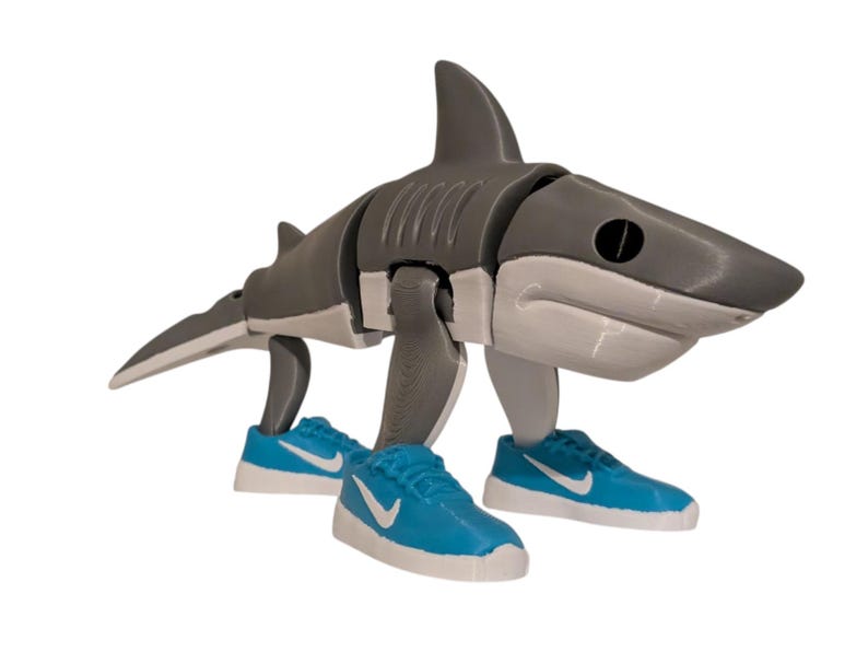May include: A gray and white articulated shark figure wearing blue sneakers with white accents. The shark has a segmented body and a fin on its back. The sneakers have a white swoosh logo.