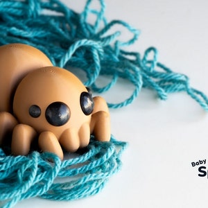 May include: A brown, cartoon-style spider toy with large black eyes, sitting on a bed of teal yarn. The text "Baby Spider" is printed below the toy.