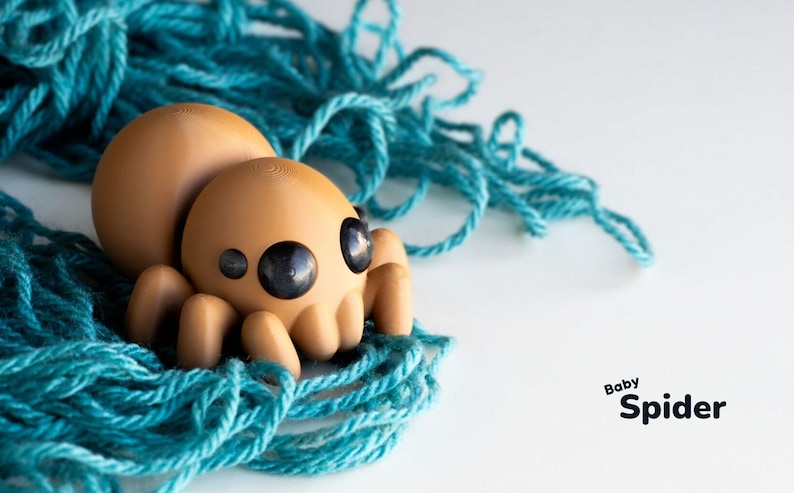 May include: A brown, cartoon-style spider toy with large black eyes, sitting on a bed of teal yarn. The text "Baby Spider" is printed below the toy.