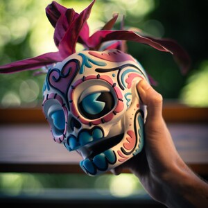 May include: A decorative skull figurine with a colorful design. The skull is painted white with blue, pink, and black accents. A dark pink plant sprouts from the top. The figurine is held in a hand.