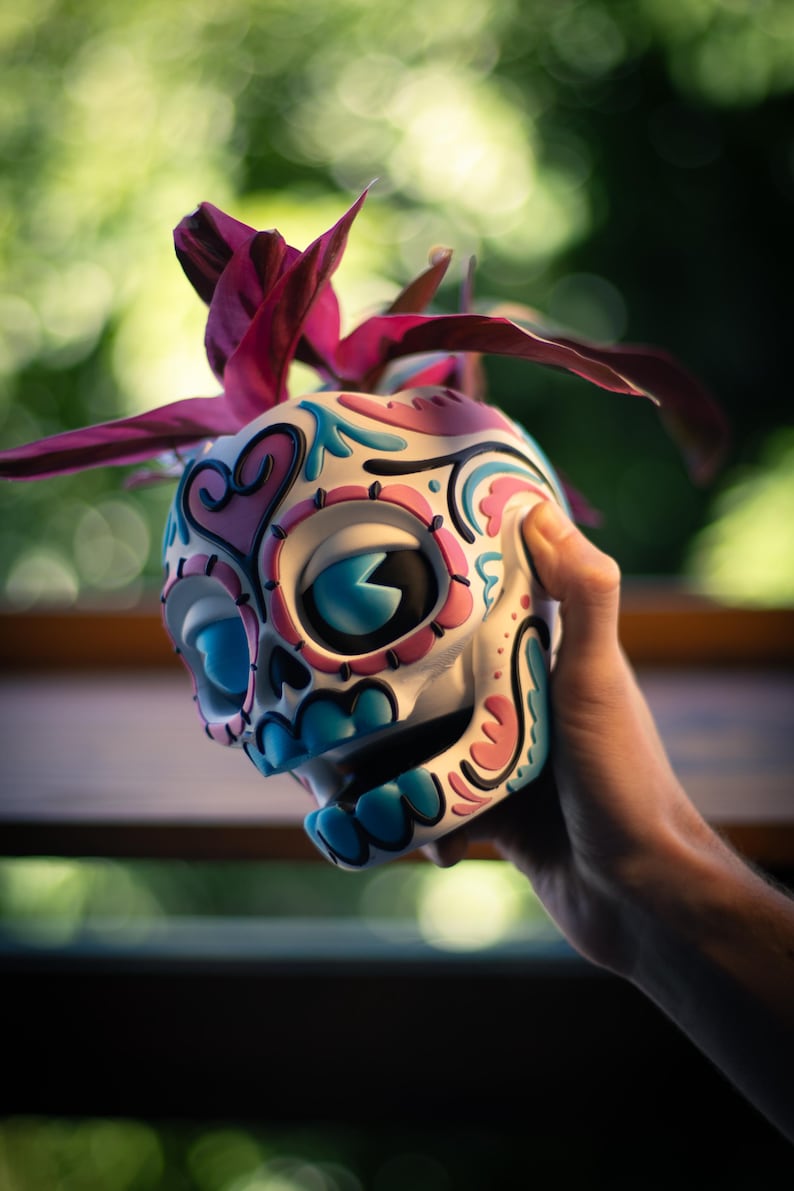 May include: A decorative skull figurine with a colorful design. The skull is painted white with blue, pink, and black accents. A dark pink plant sprouts from the top. The figurine is held in a hand.