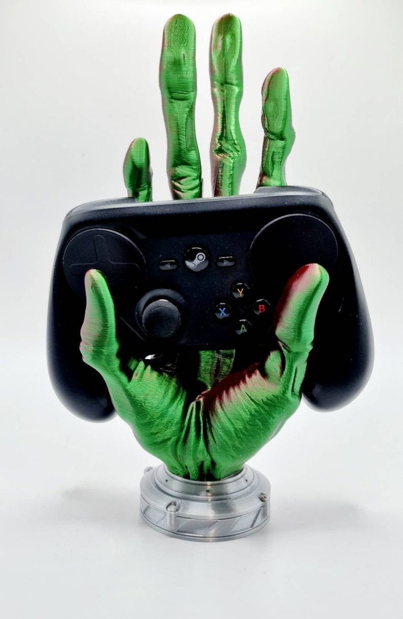 May include: A green and red skeletal hand holding a black video game controller. The hand is attached to a silver base.