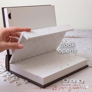 Puede incluir: Un juego de Scrabble en un estuche con forma de libro. El estuche es marr&oacute;n con p&aacute;ginas interiores blancas. La parte superior est&aacute; abierta, revelando una cuadr&iacute;cula. El texto "Book opens" es visible. Las fichas de Scrabble est&aacute;n esparcidas por la base, con las letras "B, L, E" visibles.