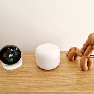 May include: A black Nest thermostat with a white base, a white Google Home Mini smart speaker, and a brown balloon animal shaped like a horse on a wooden surface.