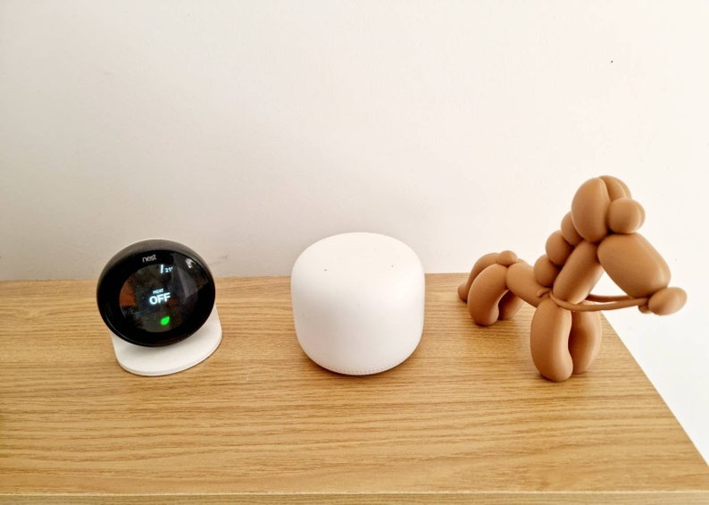 May include: A black Nest thermostat with a white base, a white Google Home Mini smart speaker, and a brown balloon animal shaped like a horse on a wooden surface.