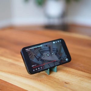 Może przedstawiać: Czarny smartfon wyświetlający wideo, podparty małym, miętowo-zielonym stojakiem. Wideo przedstawia wnętrze budynku. Telefon leży na drewnianej powierzchni. Tytuł filmu to "I Built a Secret Door to Trick my Landlord".