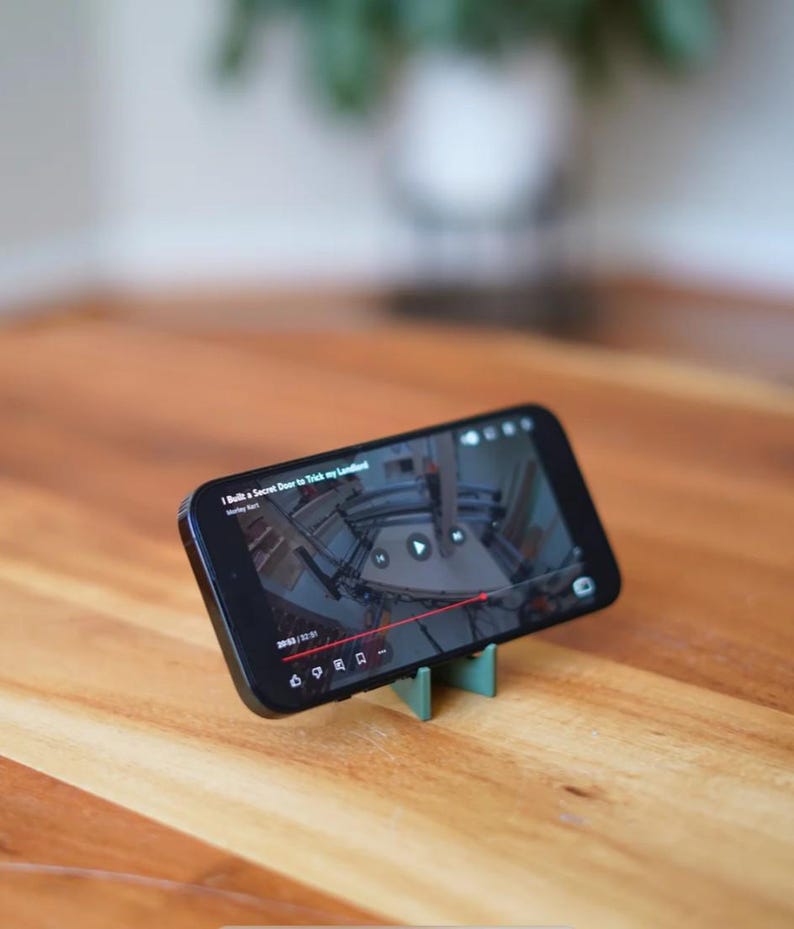 Może przedstawiać: Czarny smartfon wyświetlający wideo, podparty małym, miętowo-zielonym stojakiem. Wideo przedstawia wnętrze budynku. Telefon leży na drewnianej powierzchni. Tytuł filmu to "I Built a Secret Door to Trick my Landlord".