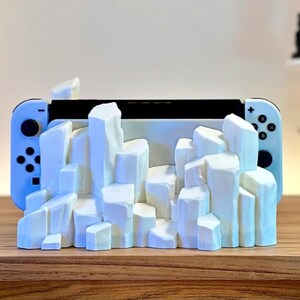 May include: A white Nintendo Switch console is displayed in a white, 3D printed stand shaped like a mountain range.