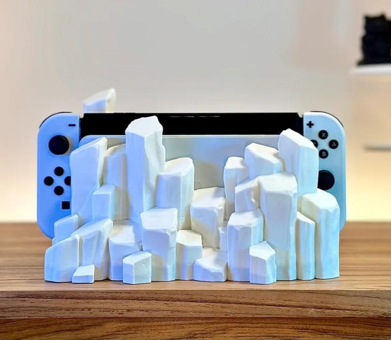May include: A white Nintendo Switch console is displayed in a white, 3D printed stand shaped like a mountain range.