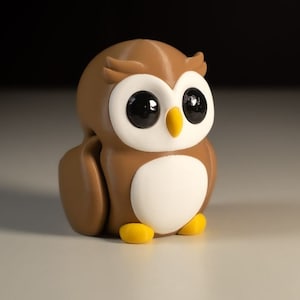 Puede incluir: Una figurita de b&uacute;ho marr&oacute;n y blanco con grandes ojos negros y patas amarillas. El b&uacute;ho est&aacute; sentado sobre una superficie blanca.