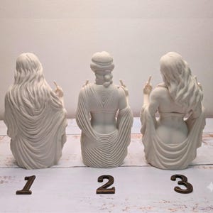 May include: Three off-white matte figurines depicting figures from the back, each with long hair or head coverings and draped clothing. Each figure is raising both middle fingers. The numbers 1, 2, and 3 are placed in front of each respective statue.