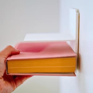 May include: A pink book with yellow-orange pages is placed on a white shelf attached to a white wall. The book is held by a hand. The shelf is a simple, modern design.