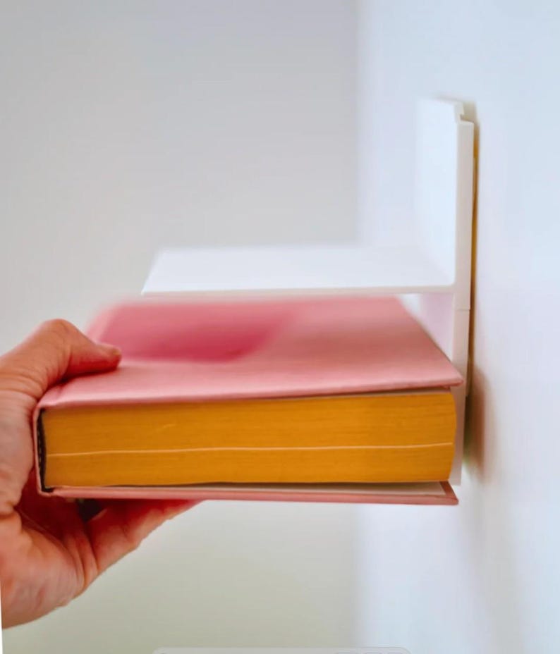 May include: A pink book with yellow-orange pages is placed on a white shelf attached to a white wall. The book is held by a hand. The shelf is a simple, modern design.