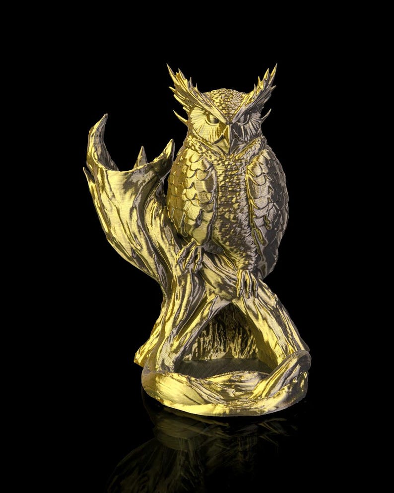 Op de afbeelding: Een gouden uil sculptuur, zittend op een tak. De uil heeft gedetailleerde veertjes en een strenge uitdrukking. De sculptuur staat tegen een zwarte achtergrond, wat de metallic afwerking en het ingewikkelde ontwerp benadrukt.