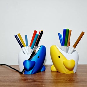 May include: Two 3D printed shark-shaped pencil holders, one blue and one yellow, with white interiors. Each holder has a variety of colored markers and pens sticking out of the top.