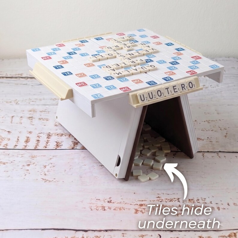 Puede incluir: Un juego de mesa Scrabble blanco con un soporte para fichas beige y un soporte de madera. El tablero tiene cuadrados azules, rojos y beige con letras que forman palabras. Las fichas adicionales se guardan debajo del tablero.