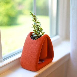 May include: A terracotta-colored, modern planter with a small succulent plant. The planter has a unique, curved design with an open base and a central pot. The plant has small green leaves and is placed on a white windowsill.
