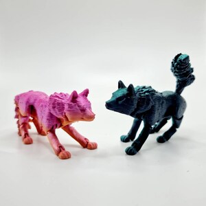 May include: Two articulated wolf figurines, one pink and one teal blue, posed facing each other on a white surface.