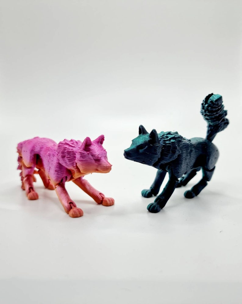 May include: Two articulated wolf figurines, one pink and one teal blue, posed facing each other on a white surface.