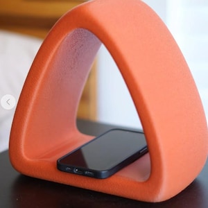 May include: An orange, triangular phone stand with a black smartphone resting inside. The stand has a smooth, textured surface and a modern design. The stand is designed to hold a smartphone.