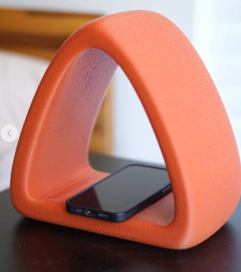 May include: An orange, triangular phone stand with a black smartphone resting inside. The stand has a smooth, textured surface and a modern design. The stand is designed to hold a smartphone.