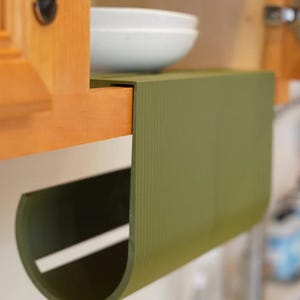 May include: An olive green, semi-circular kitchen organizer attached to a wooden cabinet. The organizer has a ribbed texture and a white horizontal stripe. A white bowl is visible above the organizer.