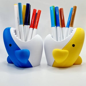 May include: Two colorful shark-shaped pencil holders, one blue and white, the other yellow and white, each filled with a variety of colored markers.