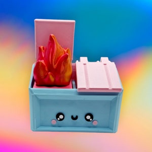 May include: A blue and pink toy dumpster with a smiling face and a red and orange flame on top. The dumpster has a lid that is open.